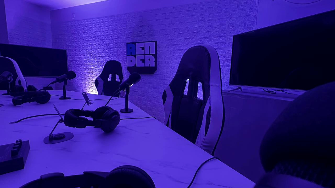 Mesa con micrófonos y auriculares, sillas y televisores, con el logo de Render en la pared